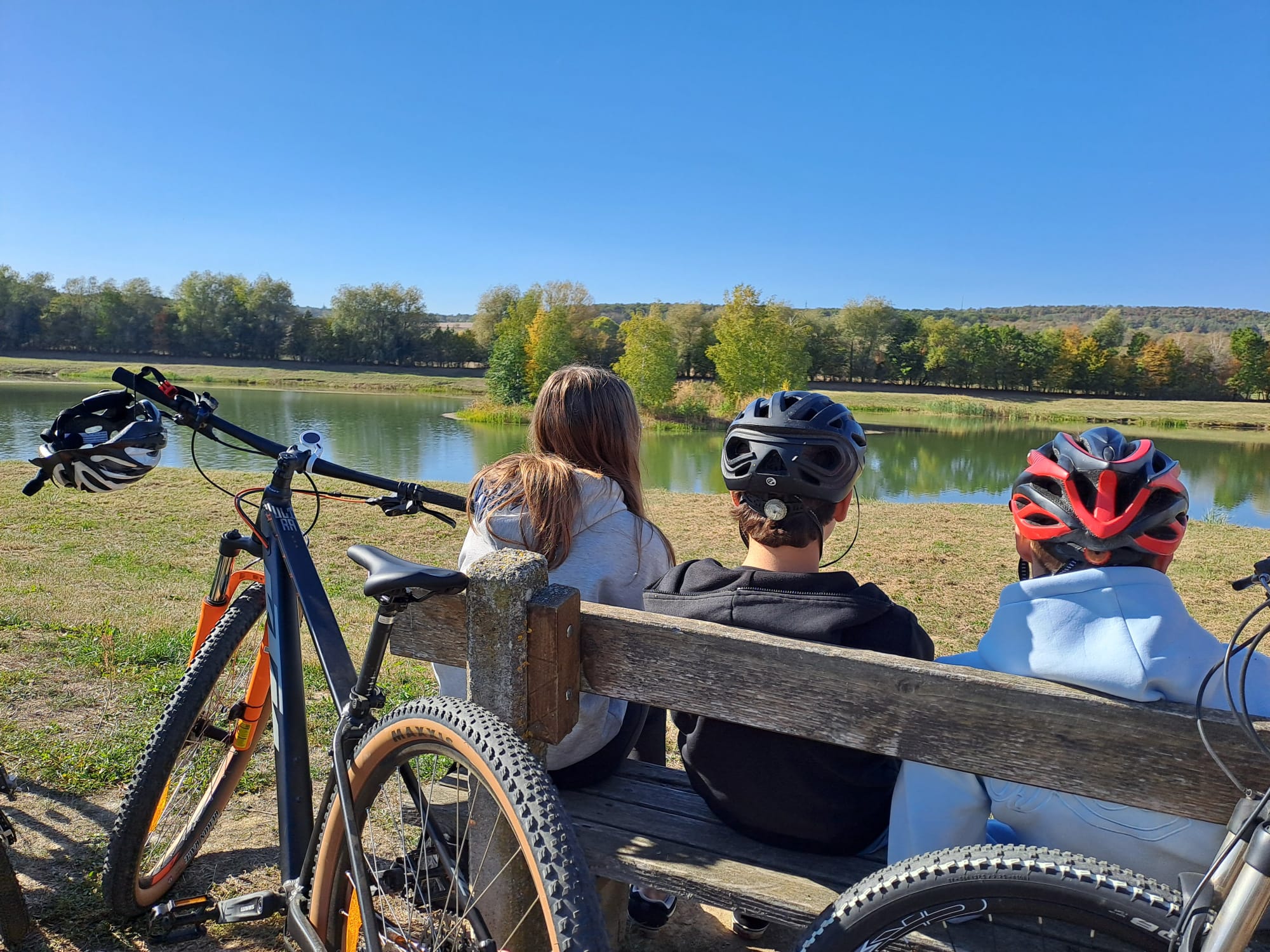 Drei Radfahrer mit Helmen sitzen auf einer Bank am See mit Bäumen im Hintergrund